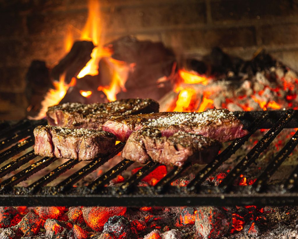 A close up of steaks on a hot grill in front of a brick backdrop